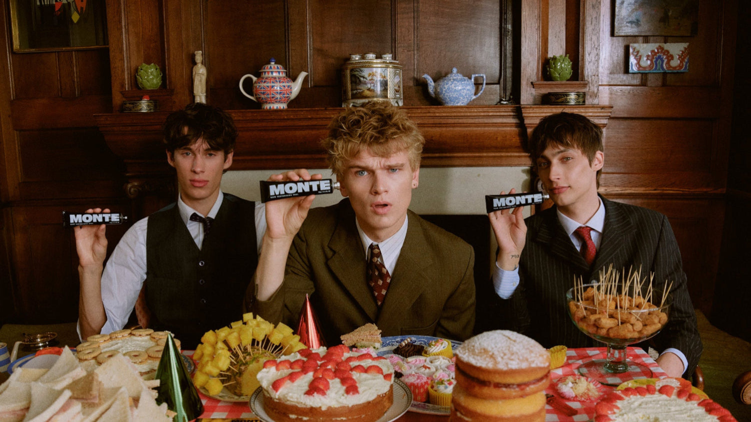 Three men sitting at a table with food and cake, holding 'Monte' signs.